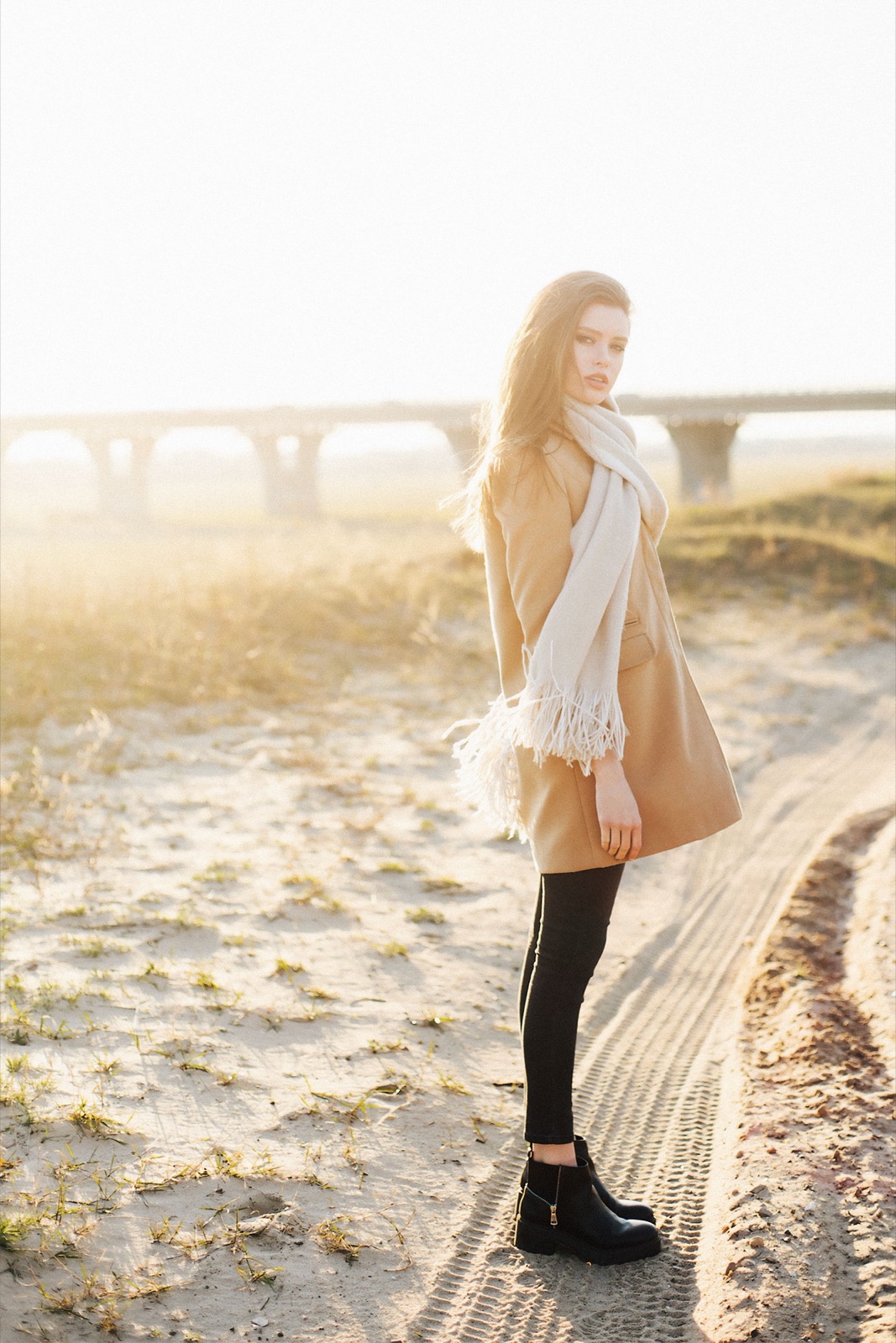 Woman wearing a camel coat, pale scarf, dark tights and ankle boots on a sunlit path.